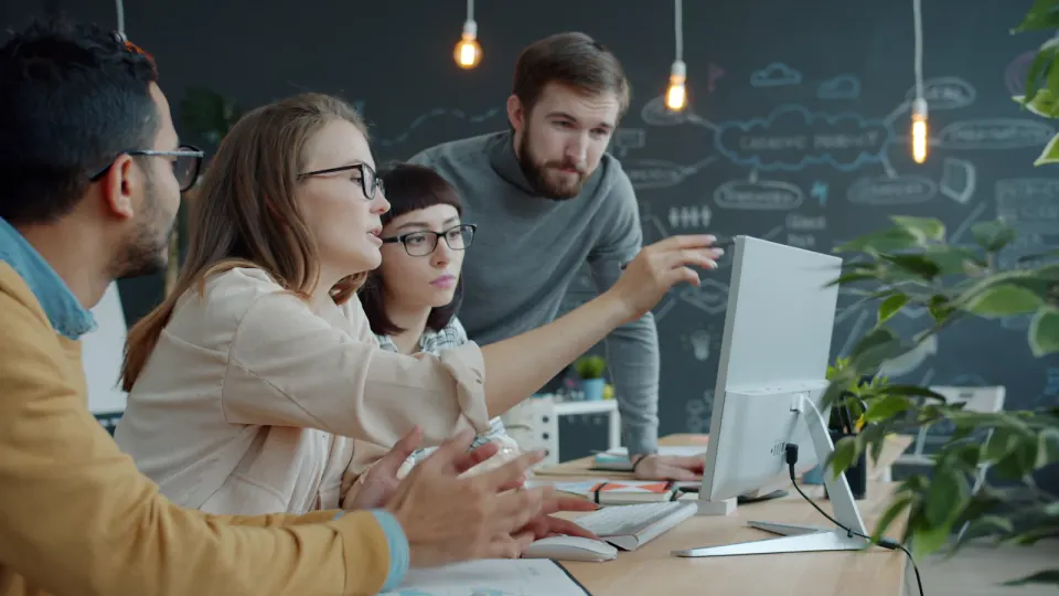 Four people discussing work around a desktop monitor, with one pointing toward the screen.