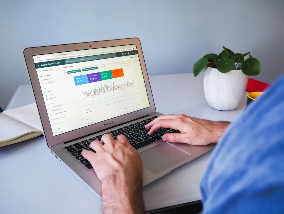 Person reviewing search-performance charts on a laptop beside a notebook and potted plant.